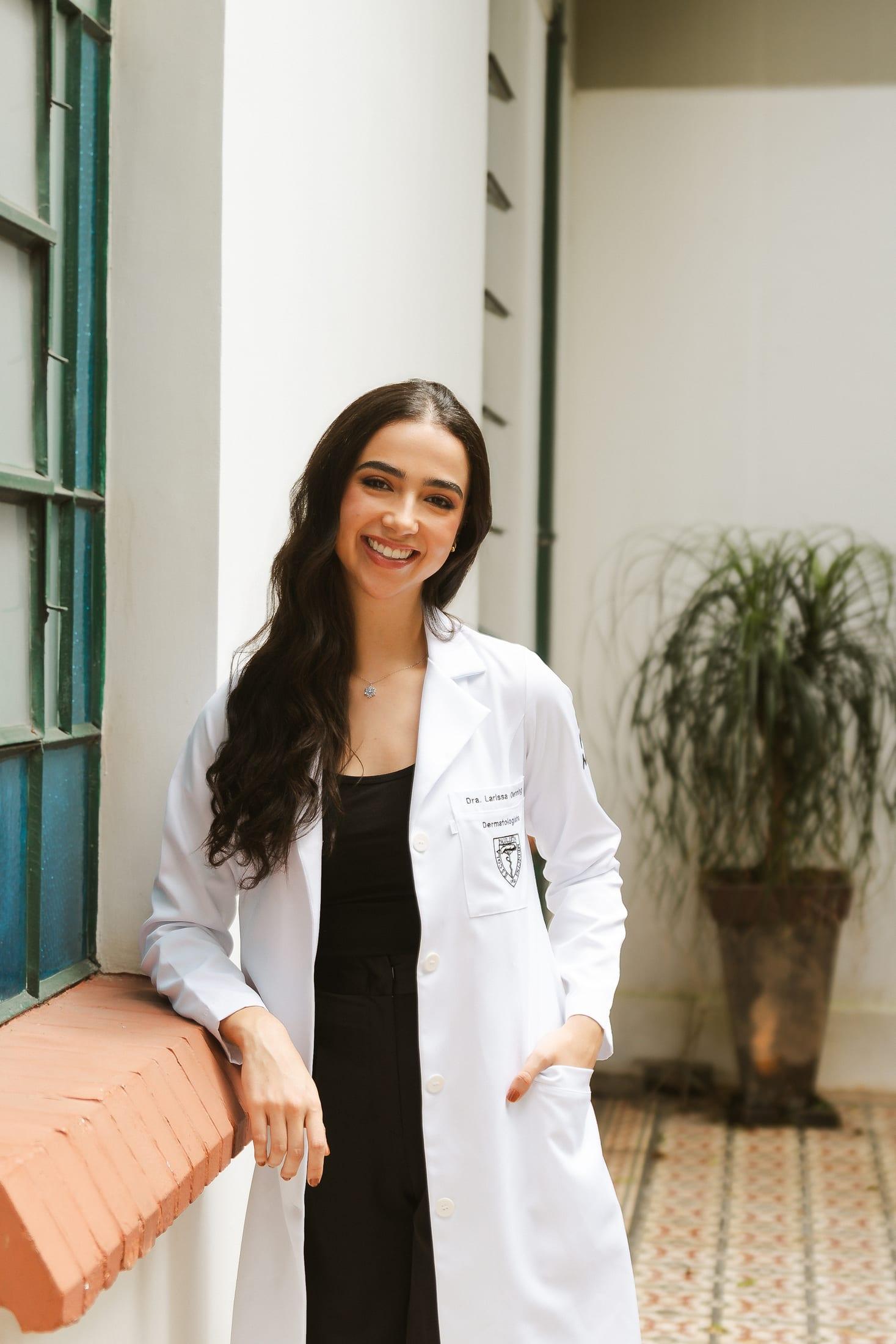 Dra. Larissa Oenning de jaleco, sorrindo, em uma varanda de arquitetura colonial